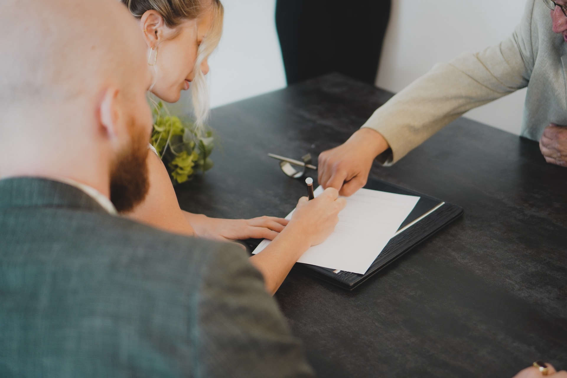 one person pointing at paperwork, one person signing paperwork, one person watching