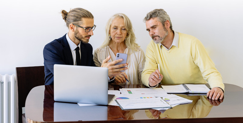 3 people looking at a cellphone. Paperwork and computer on a table.